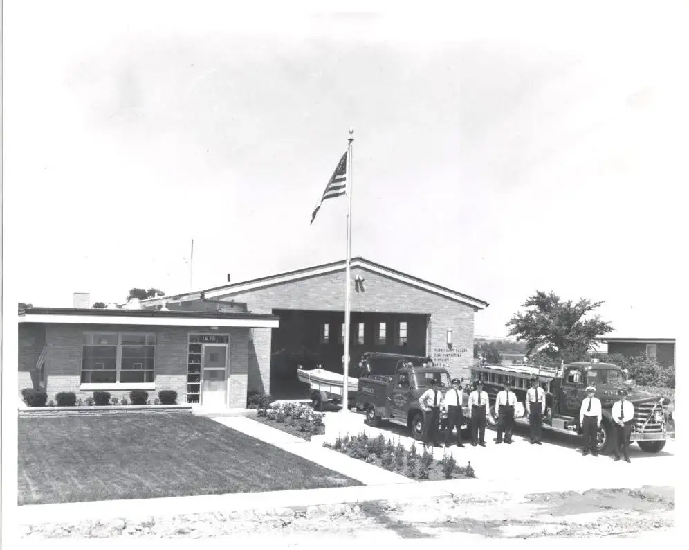 Florissant Valley firefighters lined up with apparatus and the American flag outside a mid-century brick firehouse