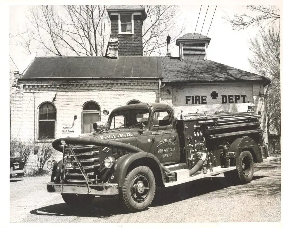 An early Florissant Valley Fire Protection District engine parked outside the original Florissant firehouse, its wall painted 'Fire Dept.'