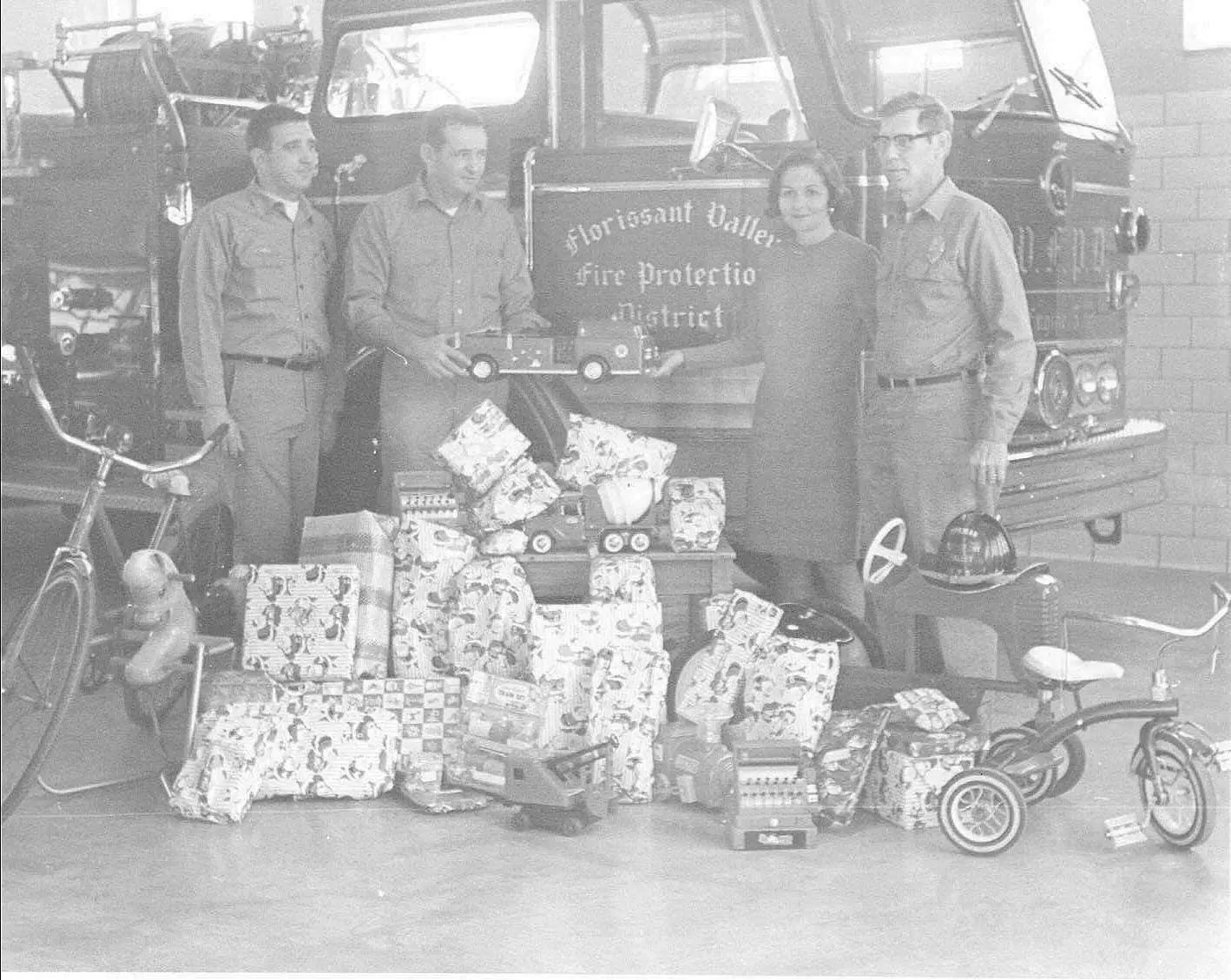 Florissant Valley firefighters and a volunteer standing with donated toys stacked in front of an engine at a holiday toy drive