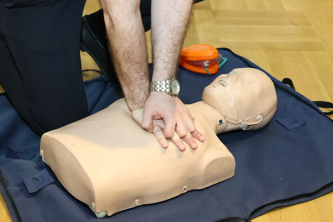 A rescuer's hands performing chest compressions on a CPR training manikin, with a training AED and pocket mask beside it